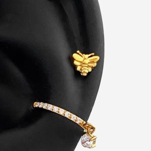 May include: A gold-toned bee stud earring and a gold-toned hoop earring featuring a row of clear, sparkling stones along the band with a single dangling clear stone charm at the end, both displayed on a black ear-shaped jewellery display form.