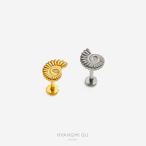 May include: Two flat-back stud earrings shaped like ammonite fossils. One earring is gold-toned with a small embedded stone, and the other is silver-toned. Both feature a spiral shell design with a post and a circular flat backing for secure wear.