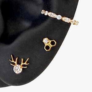 Può includere: Un set di tre orecchini color oro composto da un ear cuff curvo con pietre trasparenti, un perno esagonale a forma di nido d'ape con una pietra trasparente e un orecchino a lobo con design a corna di cervo e una pietra centrale.