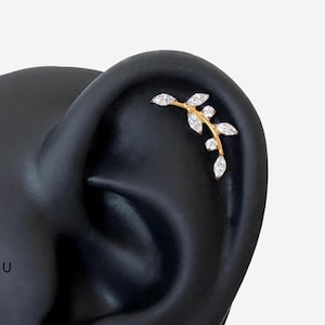 May include: A gold-toned ear climber earring shaped like a curved branch with six marquise-cut clear stones arranged as leaves. The jewelry piece follows the natural curve of the ear cartilage, featuring a polished metal stem and sparkling stones.