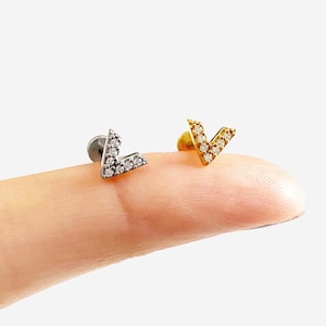 May include: Two V-shaped stud earrings, one silver and one gold, each set with small, clear gemstones. The earrings are shown against a light background, highlighting their delicate design and sparkle.