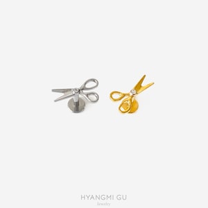 May include: Two flat-back labret studs featuring scissor-shaped charms. One stud is silver-toned and the other is gold-toned. Each scissor charm has a small, clear, round-cut stone set at the pivot point. The text HYANGMI GU Jewelry is at the bottom.