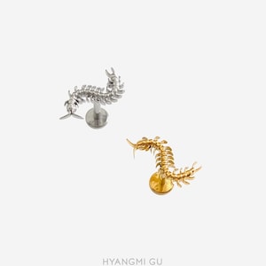 May include: Two body jewellery pieces featuring a centipede design, one in a silver tone and one in a gold tone. Each piece has a flat circular base and a post, with the detailed centipede figure attached to the top, positioned in a curved shape.