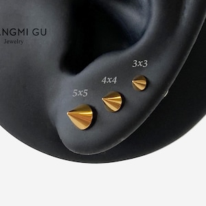 Puede incluir: Tres pendientes de botón dorados en forma de cono dispuestos en fila sobre un modelo de oreja oscuro. Los pendientes están etiquetados con texto que indica los tamaños 5x5, 4x4 y 3x3. El texto NGMI GU Jewelry es visible arriba a la izquierda.