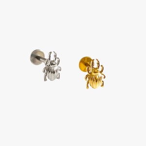 May include: Two flat back stud earrings shaped like beetles. One earring is silver-toned metal, and the other is gold-toned metal. Each stud features a detailed insect design with prominent mandibles and a rounded body, attached to a circular post.