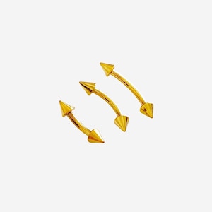 May include: Three gold-colored curved eyebrow rings with pointed ends. The jewelry is set against a white background. The curved bars have a slight arch, and the pointed ends are conical.