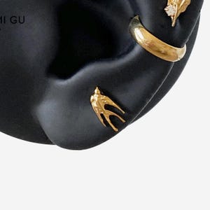 May include: A gold-toned swallow stud earring positioned below a smooth gold-toned hoop earring and a small gold-toned stud earring with a clear stone, all displayed on a black ear-shaped stand with partial text visible in the upper left corner.