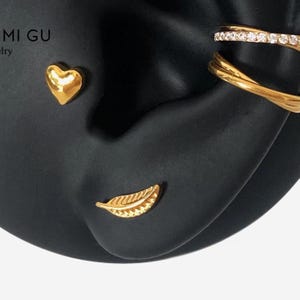 Könnte beinhalten: Ein Set aus goldfarbenem Schmuck, bestehend aus einem herzförmigen Ohrstecker, einem blattförmigen Ohrstecker und einer mehrreihigen Ohrklemme mit einer Reihe kleiner klarer Steine. Der Schmuck ist auf einem schwarzen Ohr-Modell mit Text MI GU.