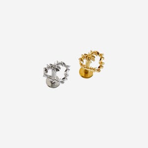 May include: Two bottle stoppers, one silver and one gold. Each has a circular design of small, leaf-like shapes. The stoppers have a round base. The background is white. The stoppers are designed to fit into the neck of a bottle.