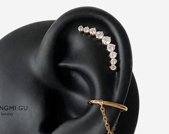 Può includere: Un orecchino a lobo dorato con una fila curva di sette pietre rotonde trasparenti posizionate lungo la cartilagine superiore, abbinato a un ear cuff dorato con un delicato design a catenella pendente sul lobo inferiore.