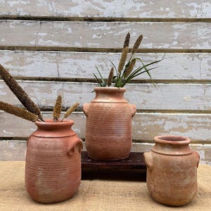 May include: Three terracotta vases of varying sizes, each with a rustic, textured finish and small handles. The vases are filled with dried grasses and sit on a burlap surface against a weathered white wall.