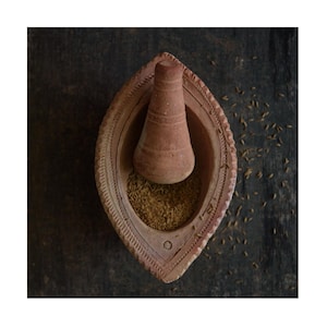 May include: A terracotta mortar and pestle with a brown wooden background. The mortar is filled with ground cumin seeds.