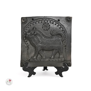Könnte beinhalten: Eine dunkelgraue, quadratische Fliese mit einem detaillierten Relief eines Tigers in einem dekorativen Rahmen. Die Fliese wird von einem schwarzen Metallständer getragen. Der Tiger hat ein gestreiftes Muster und ist von floralen und geometrischen Designs umgeben.