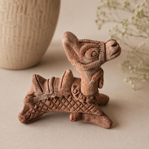 May include: A small, hand-sculpted terracotta figurine of a camel in a resting position. The figure features detailed etched patterns on its body, a textured saddle, and a harness around its head, all rendered in a natural, earthy reddish-brown clay.