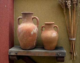 May include: Two terracotta vases with handles sit on a rustic wooden shelf. The vases are different sizes and have a weathered appearance. A bundle of wheat stalks is tied with twine and leans against the wall, adding a natural element to the display.