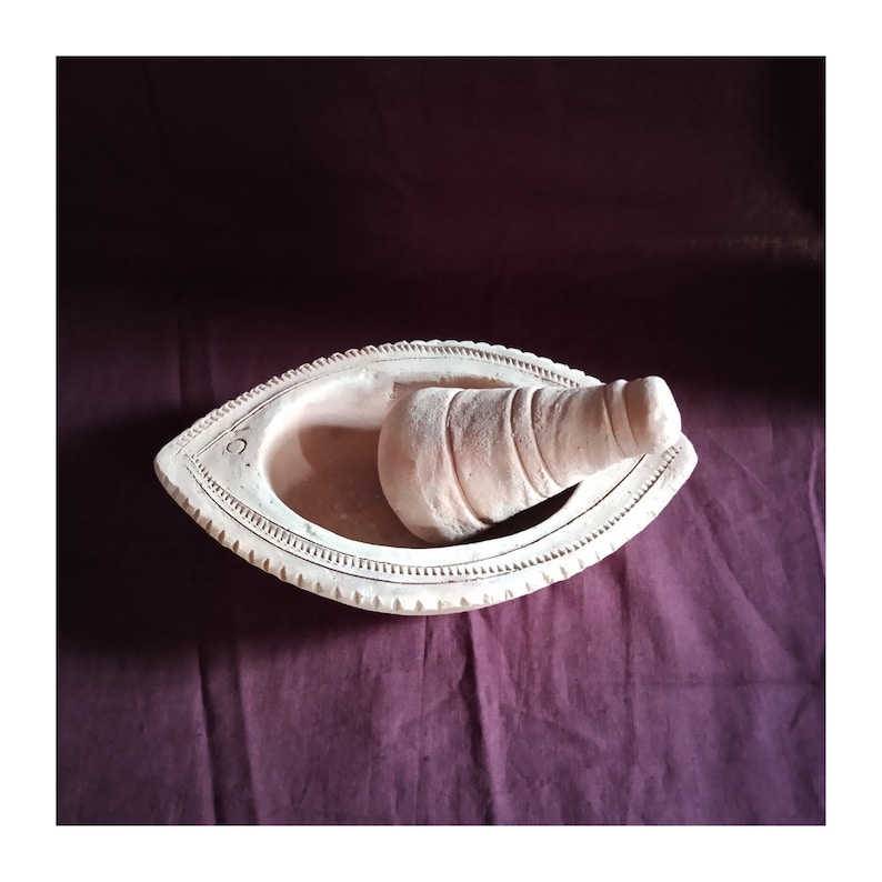 May include: A terracotta mortar and pestle set. The mortar is shaped like a boat and has a decorative design around the rim. The pestle is a cylindrical shape with a rounded end.