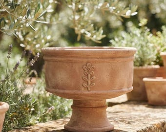 May include: A terracotta planter with a wide, circular bowl and a pedestal base. The exterior features a raised, embossed flower design on the front. The surface has a natural, matte, earthy orange finish with subtle variations in texture.