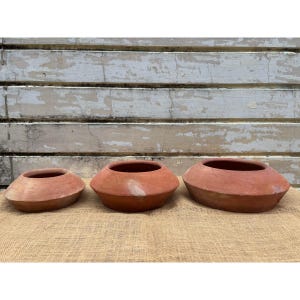 Pode incluir: Três vasos redondos rasos de cor terracota, de tamanhos variados. Os vasos têm uma borda larga e angulada e uma superfície lisa e sem vidrado. Estão dispostos sobre uma superfície de serapilheira contra um fundo de madeira pintada de branco desgastado.