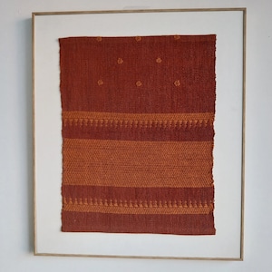 Op de afbeelding: Een ingelijst geweven wandkleed met een bruin en oranje geometrisch patroon. Het wandkleed heeft een textuur en is ingelijst in een licht houten frame.