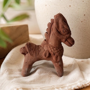 May include: A small, hand-sculpted terracotta horse figurine with a matte, reddish-brown finish. The horse features detailed carvings on its mane, a textured saddle area, and a stylized head with defined eyes and a bridle-like pattern across its face.