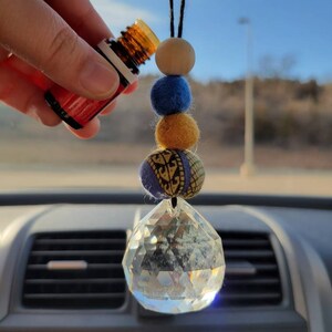 May include: A crystal car charm with a beaded string hanging from a rearview mirror. The beads are blue, yellow, and wood. A hand is holding a small bottle of essential oil over the charm.