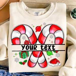 May include: A cream-colored sweatshirt with a Christmas-themed design featuring two red and white candy canes, a red ornament, a green gift box, a green holly sprig, a red Santa hat, and snowflakes. The text "YOUR TEXT" is printed on a banner between the candy canes.