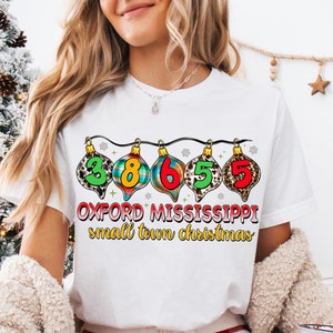 Może przedstawiać: Biała koszulka z nadrukiem świątecznej ozdoby z numerami 3865 i tekstem "Oxford Mississippi small town christmas".