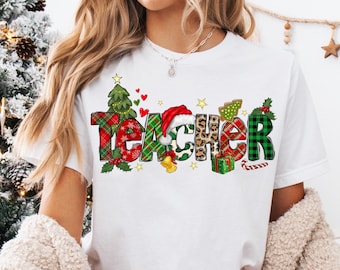 Puede incluir: Una camiseta blanca con un diseño navideño que dice "Teacher" en letras de colores a cuadros y estampadas. Las letras están decoradas con árboles de Navidad, adornos y bastones de caramelo.