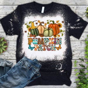 Puede incluir: Una camiseta negra con un dise&ntilde;o salpicado de lej&iacute;a que presenta un gr&aacute;fico colorido de "Pumpkin Patch" con calabazas, calabacines y mariposas.