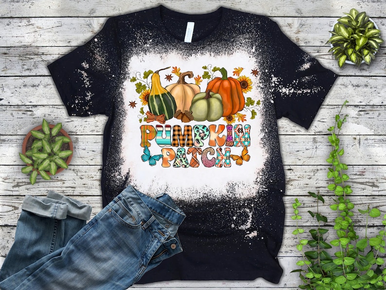 Puede incluir: Una camiseta negra con un dise&ntilde;o salpicado de lej&iacute;a que presenta un gr&aacute;fico colorido de "Pumpkin Patch" con calabazas, calabacines y mariposas.