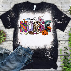 Pu&ograve; includere: Una maglietta nera con un design spruzzato di candeggina che presenta la parola "NURSE" in lettere colorate con accenti a tema Halloween, tra cui un cappello da strega, una scopa e zucche.