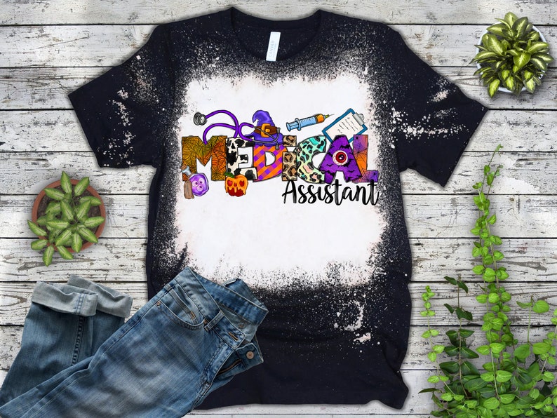 May include: A black t-shirt with a white bleach design featuring the words "Medical Assistant" with Halloween-themed letters and icons.
