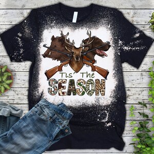 Peut inclure: Un t-shirt noir avec un motif blanchi repr&eacute;sentant un cerf, un sanglier et un chien avec des fusils crois&eacute;s et le texte "Tis' The Season".