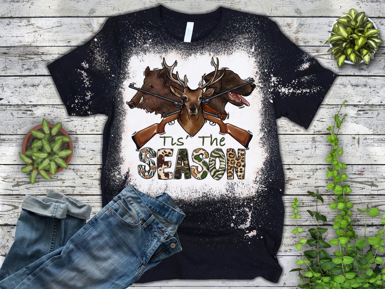 Peut inclure: Un t-shirt noir avec un motif blanchi repr&eacute;sentant un cerf, un sanglier et un chien avec des fusils crois&eacute;s et le texte "Tis' The Season".
