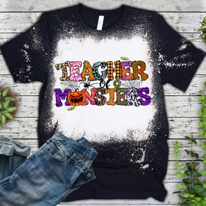 May include: Black t-shirt with a bleach-splattered design. The shirt features a colorful graphic that says "Teacher of Monsters" with a spider and a pumpkin.