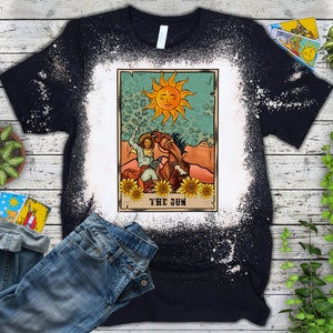 May include: Black t-shirt with a bleached white design. The graphic features a tarot card illustration with a sun, a person on a horse, and the text "THE SUN". Sunflowers and a leopard print background complete the design.