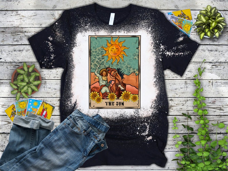 May include: Black t-shirt with a bleached white design. The graphic features a tarot card illustration with a sun, a person on a horse, and the text "THE SUN". Sunflowers and a leopard print background complete the design.