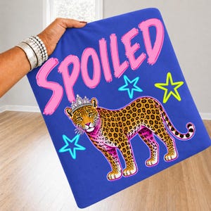 Op de afbeelding: Een blauwe deken met het woord "SPOILED" in roze en een luipaard met een kroon. De luipaard heeft een roze en geel ontwerp, met blauwe en gele sterren. De deken wordt door een persoon vastgehouden.