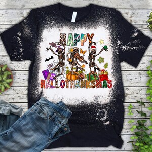 May include: A black t-shirt with a bleach-splattered design featuring three skeletons dancing with the text "Happy Hallothanksmas".