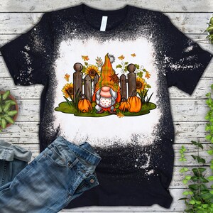 Op de afbeelding: Een zwart T-shirt met een bleekmiddelgespat design met een gnome met een rood-witte muts en een witte baard. De gnome staat voor een hek met pompoenen en zonnebloemen.