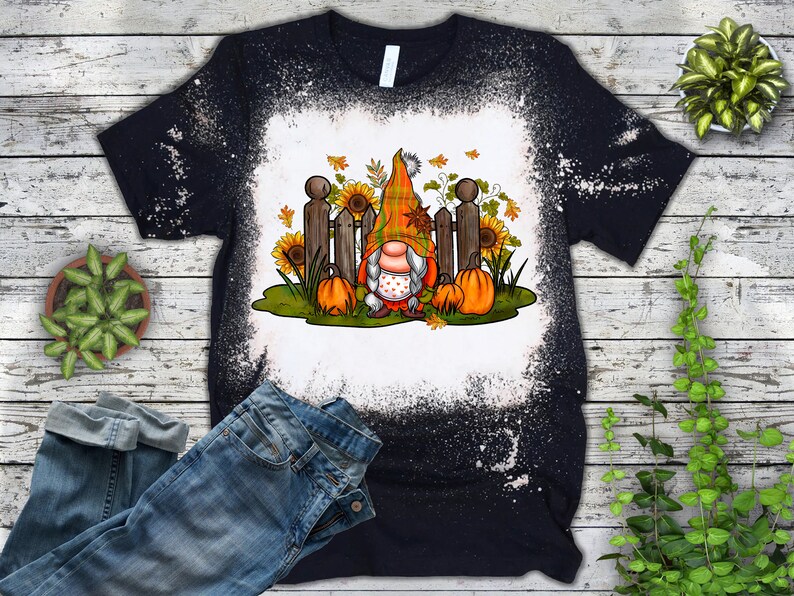 Op de afbeelding: Een zwart T-shirt met een bleekmiddelgespat design met een gnome met een rood-witte muts en een witte baard. De gnome staat voor een hek met pompoenen en zonnebloemen.