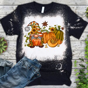 May include: A black t-shirt with a bleach-splattered design featuring a cartoon gnome holding a pumpkin. The gnome is wearing a red and orange hat with sunflowers. The design also includes pumpkins and autumn leaves.