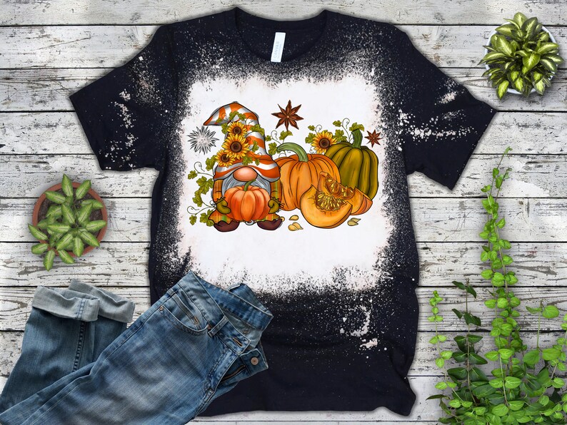 May include: A black t-shirt with a bleach-splattered design featuring a cartoon gnome holding a pumpkin. The gnome is wearing a red and orange hat with sunflowers. The design also includes pumpkins and autumn leaves.