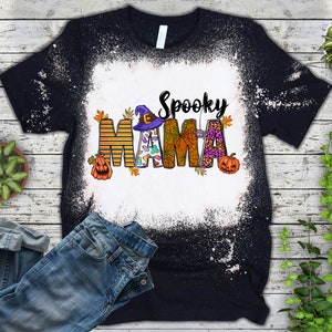 Pode incluir: Camiseta preta com um centro branco descolorido, com as palavras "Spooky Mama" em um design tem&aacute;tico de Halloween. O design inclui um chap&eacute;u de bruxa, ab&oacute;boras e folhas de outono. Uma cal&ccedil;a jeans azul &eacute; vis&iacute;vel na imagem.