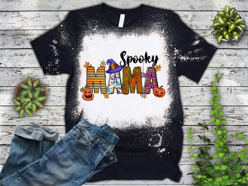 Pode incluir: Camiseta preta com um centro branco descolorido, com as palavras "Spooky Mama" em um design tem&aacute;tico de Halloween. O design inclui um chap&eacute;u de bruxa, ab&oacute;boras e folhas de outono. Uma cal&ccedil;a jeans azul &eacute; vis&iacute;vel na imagem.