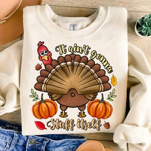 May include: A white sweatshirt with a Thanksgiving-themed design. A cartoon turkey with a red head and brown feathers is depicted with the text "It ain't gonna stuff itself".