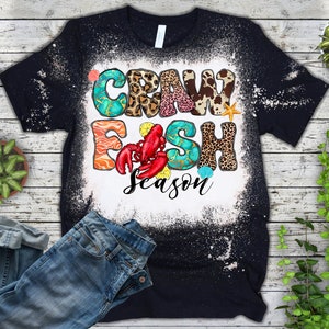 Peut inclure: Un t-shirt noir avec un motif &eacute;clabouss&eacute; d'eau de Javel repr&eacute;sentant le texte "Crawfish Season" en lettres color&eacute;es avec des accents d'imprim&eacute;s animaliers et de coquillages.