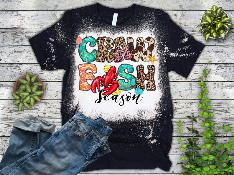 Peut inclure: Un t-shirt noir avec un motif &eacute;clabouss&eacute; d'eau de Javel repr&eacute;sentant le texte "Crawfish Season" en lettres color&eacute;es avec des accents d'imprim&eacute;s animaliers et de coquillages.