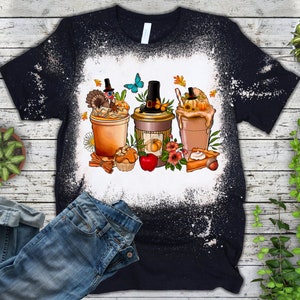 Puede incluir: Una camiseta negra con un dise&ntilde;o de blanqueador blanco de tres bebidas con tem&aacute;tica oto&ntilde;al con un pavo, una calabaza y un pastel en la parte superior. El dise&ntilde;o tambi&eacute;n incluye hojas de oto&ntilde;o, una mariposa y palitos de canela.