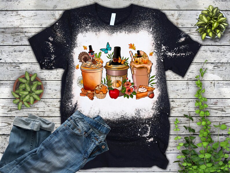 Puede incluir: Una camiseta negra con un dise&ntilde;o de blanqueador blanco de tres bebidas con tem&aacute;tica oto&ntilde;al con un pavo, una calabaza y un pastel en la parte superior. El dise&ntilde;o tambi&eacute;n incluye hojas de oto&ntilde;o, una mariposa y palitos de canela.